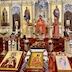 View of the Fr. Aleksey standing at a podium in front of the opening archway for the Royal Doors. There are altar servers on either side of him, and each is holding a large candle.