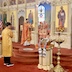 Archbishop Mark stands in the archway of the open Royal Doors holding up a covered chalice towards the congregation. Fr. Aleksey is standing to his right, with another chalice that is similarly covered by an orange and gold cloth. The cloth match the clergy's vestments. There's also an altar server at the foot of the three steps that lead up to the archway. The alter server is facing Archbishop Mark and is wearing a gold vestment and holding a large candle in a gold candle holder.