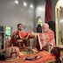 Fr. Aleksey and Archbishop Mark are standing at the altar table, holding an orange and gold cloth over a chalice and bread during preparation of communion. The archbishop's mitre appears on the table in the left-bottom corner of the frame, and a few parishoners who are seated in pews in the body of the church are visible through the open archway along the right-hand-side of the frame.