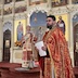 Father Aleksey is standing at the front of the church, speaking to the congregation, and holding a piece of paper in his hands. Fr. Aleksey is in sharp focus, appearing in the right-hand third of the frame, while Archbishop Mark, who is slightly out of focus, appears in the center of the photo. The archbishop is standing in front of the open Royal Doors and is holding a sliver cross in his right hand, and a crozier in his left hand. He's looking in the direction of Fr. Aleksey and listening.