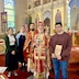 A portrait photo of Katerina, Matushka Natallia, Father Aleksey, Archbishop Mark, and Michael. They're posing in front of the left front corner of the body of the church, and the iconostas appears across the right two thirds of the background, with a stained glass window and its corresponding wall appearing on the left third of the background. There's an American flag in the corner, between the wall and iconostas. Archbishop Mark is holding a cross in his right hand, and his crozier in his left hand; he's also wearing his mitre. Michael is holding an icon of St. Michael the Archangel..