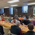 Parishoners sitting at tables in the church hall while eating and talking. There are two parallel tables that are covered by orangey-gold table cloths and that run at a diagonal from the lower right corner to the center of the left frame.