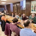 A table, covered in an orangey-gold tablecloth, in the church hall, where parishoners are seated while eating and talking. The table runs from the lower right corner to the middle of the left side of the frame.