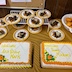 Two cakes and slices of cake on a table that is covered with a gold table cloth. The half of the table closest to the view has two cakes with white icing and yellow writing. The cake on the left says Welcome Arch Bishop Mark, and the cake on the right says Welcome Home Michael. Both cakes have an orange rose with smaller yellow roses and green leaves in the lower left corner of the cake. Both also have orange trim around the edges of the cake. The back half of the table has places with chocolate cake slices with a light brown icing (looks like a maple icing color but may not be maple-flavored). There's a white collander filled with Mandarin oranges to the right of the cake slices, and it appears almost in the upper right corner of the photo.