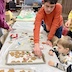 A few people standing on either side of a table where a male is placing placing a baked cookie on a parchment-covered baking sheet with other cookies in preparation for decorating. A young boy is seated and watching, while a woman is standing across the table working and another woman is partially visible behind the boy.