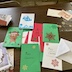 A variety of homemade Christmas cards on a brown table. The cards are mostly red, green, or white with blue, and there are sheets with a few remaining festive stickers that were placed on the cards.