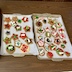 Three baking sheet pans of colorul cookies on a table. There's parchment paper between the cookies and the baking pans, and the cookies are shaped in circles, stars, and trees, and they're decorated in various combinations of icing, M&Ms, and gel candies.